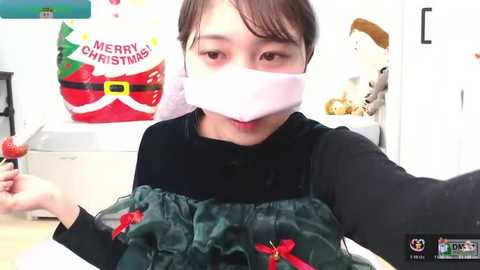 A video of an Asian woman in a black mask, wearing a festive green dress with red bows, holding a strawberry. Background features a \"Merry Christmas\" banner and stuffed animals.