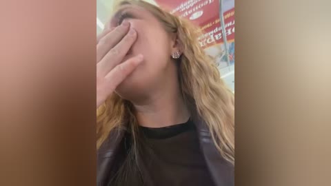 Media: Video of a blonde woman with wavy hair, wearing a black leather jacket, covering her mouth with her hand, in a blurred background with red and white signage.