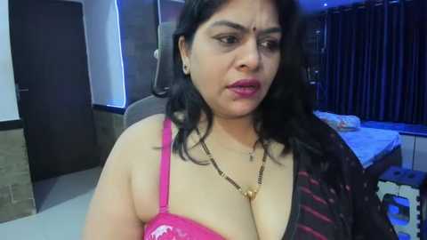 Media: Video of a middle-aged Indian woman with long black hair, wearing a pink bra, necklace, and black shawl, sitting on a bed with dark curtains in a dimly lit room.