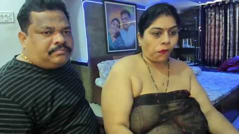 Media: Video of an Indian couple sitting on a bed in a modern bedroom. The man, with a mustache, wears a striped shirt. The woman, with dark hair and red lipstick, holds a black shawl, revealing her bare chest.