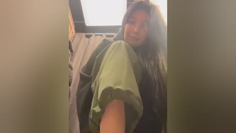 Media: Video of a young woman with long, dark hair in a bathroom. She wears a light green shirt and dark pants, leaning against a beige curtain with a window above.