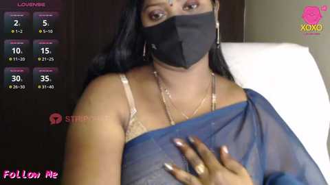 Media: Video of a South Asian woman with medium skin tone, wearing a blue saree, black face mask, and gold jewelry, in a hospital bed, with a COVID-19 tracker screen in the background.