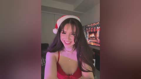 Media: Video of a smiling, slender woman with long dark hair, wearing a red lace bra and Santa hat, in a dimly lit living room with a TV displaying a fireplace scene.