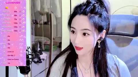 Media: Video of a fair-skinned, dark-haired Asian woman with red lipstick, wearing a blue blouse, sitting indoors near a microphone. Background shows a closet and a blue chair.