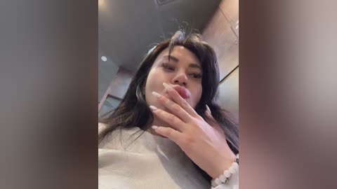 Media: Video of a young woman with long, dark hair, wearing a white sweater, playfully licking her fingers, in a blurred indoor setting.