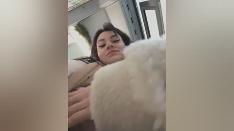 Media: Video of a woman with long dark hair, lying on a bed, wearing a light brown sweater, cuddling a white teddy bear. The background includes a mirror and a green plant.