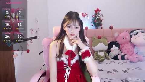 Media: Video of an East Asian woman in a red, fur-trimmed Santa outfit, sitting on a pink gaming chair in a cozy, festive bedroom with Christmas decorations.