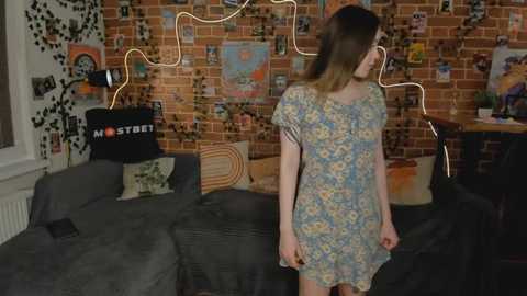 Media: Video of a young woman with long brown hair in a floral dress, standing in a cozy living room with exposed brick wall, eclectic decor, and string lights.