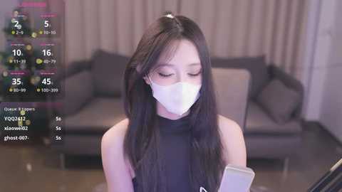 Media: Video of an East Asian woman in a black turtleneck, white mask, and long black hair, wearing a smartwatch, sitting in a modern living room with a grey couch.