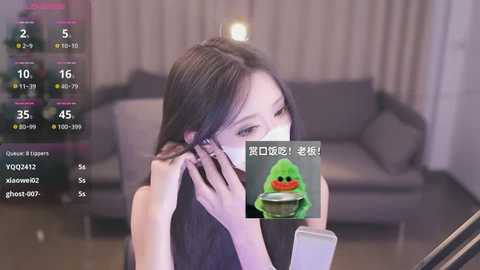 Media: A video of an East Asian woman with long black hair, wearing a face mask, speaking on a smartphone in a modern living room.