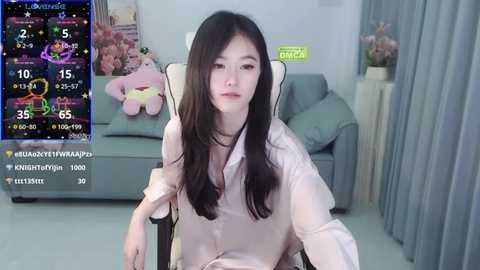 Media: Video of a young East Asian woman with long black hair, wearing a pink blouse, seated on a gray chair in a modern living room with a TV showing a colorful video game screen.