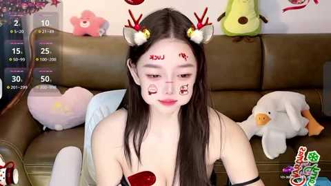 Media: Video of a young East Asian woman with long dark hair, wearing red reindeer antlers, festive makeup, and a black bra, surrounded by plush toys on a brown couch.