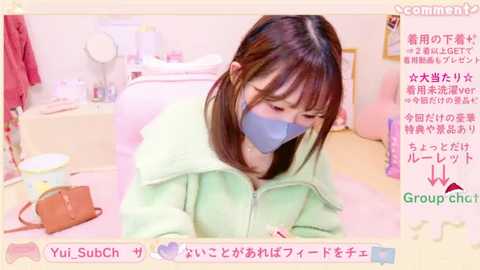 A video of a young woman with straight brown hair wearing a light green hoodie and a blue mask, sitting at a table in a cozy, pastel-colored room.