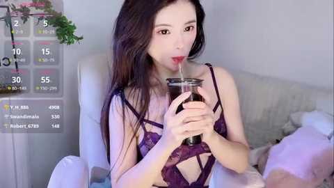 Media: Video of a young Asian woman with long black hair, wearing purple lace lingerie, drinking from a glass, seated on a white couch, with a digital calendar in the background.