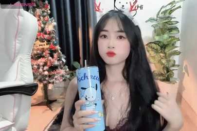 Media: Video of an East Asian woman with long black hair, red lipstick, and a black lace bra, holding a blue can featuring a cat, in a cozy indoor setting with a Christmas tree and potted plants.