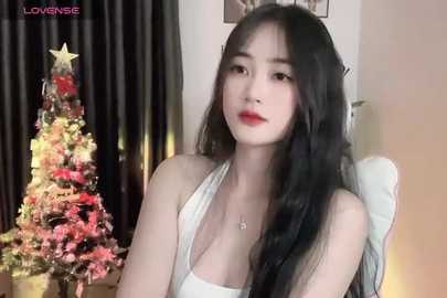 Media: A video of a young East Asian woman with long black hair, fair skin, and red lipstick, wearing a white halter top, sitting in front of a lit Christmas tree in a cozy room.