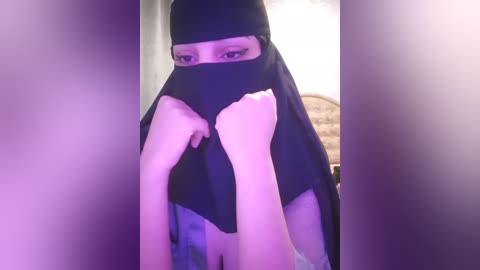 Media: Video of a person wearing a black niqab, covering the face except for the eyes, with light skin tone and medium-sized breasts partially visible. Background shows a beige wall and a bed.