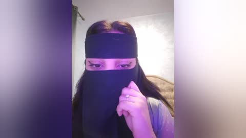 Media: Video of a woman with long dark hair, wearing a black niqab covering her face, holding it up with her right hand. She has a light complexion and is indoors, with a beige wall in the background.