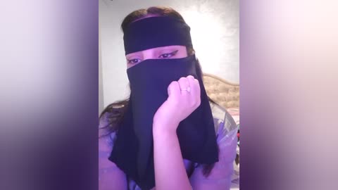 Media: Video of a woman with long brown hair, wearing a black hijab, covering her mouth with one hand. She has light skin and is indoors, with a blurred background showing a beige wall and a wooden headboard.