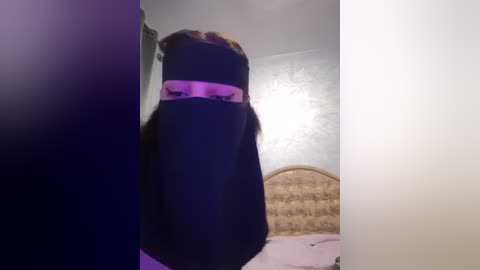 Media: A video of a person in a black face mask with long brown hair, standing in a dimly lit bedroom with a patterned headboard and lavender walls.