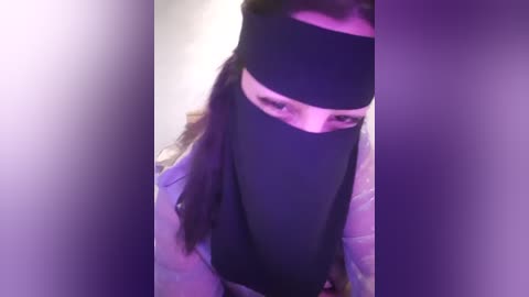 Media: Video of a person with long brown hair, wearing a black headband with a white stripe, and a black hijab, standing against a purple backdrop, blurry and slightly out of focus.