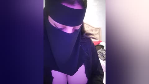 Media: A video of a person wearing a black niqab, headscarf, and jacket in a dimly lit room with a bed and scattered clothes in the background.