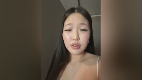 Media: A video of an Asian woman with long black hair, light skin, and neutral expression, standing in a dimly lit room with brown walls and a ceiling.