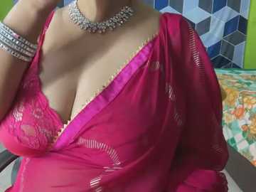 Media: Video of a woman in a pink saree, revealing a purple lace bra, adorned with silver jewelry. Background features a colorful quilt and geometric wall decor.