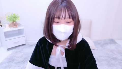 Media: Video of an East Asian woman with shoulder-length brown hair, wearing a black velvet cape, white mask, and white shirt, sitting on a grey carpeted floor, next to a white bedside table with a green plant.