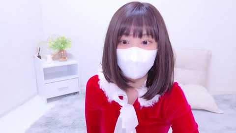 Media: Video of a young Asian woman with straight black hair, wearing a red Santa costume with white fur trim and a white mask, in a minimalist white room with a potted plant and a bed.