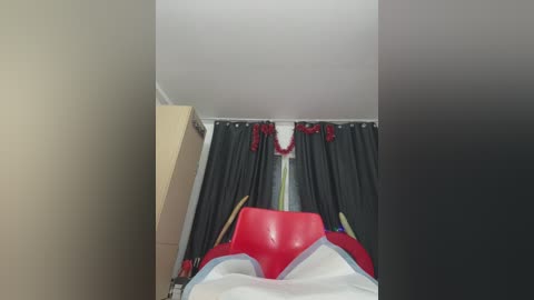 Media: Video of a simple bedroom with a red chair, dark curtains, a white wall, and a light wooden cabinet. The scene is dimly lit, giving a cozy, understated ambiance.