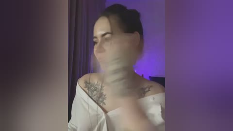 Media: Video of a woman with fair skin, dark hair in a bun, wearing an off-shoulder white top, revealing a detailed floral tattoo on her right shoulder. Background is dimly lit with purple and blue hues.