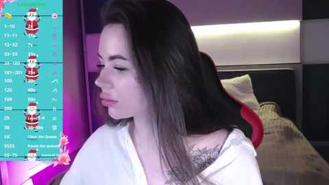 Media: Video of a young woman with long dark hair, wearing a white hoodie, sitting on a red gaming chair in a dimly lit bedroom. A video game overlay with stats is visible on the left.