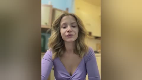 Media: A video of a fair-skinned woman with wavy blonde hair, wearing a lavender V-neck top, standing in a kitchen with yellow cabinets and blue walls. She has a serene expression.