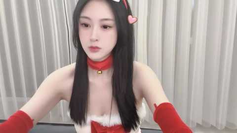 Media: Video of an East Asian woman with fair skin, long black hair, and red choker with bell. She wears a red Santa costume with white fur trim, red gloves, and a heart headband, sitting in front of white vertical blinds.