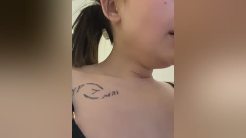 Media: A close-up video of a person's shoulder and neck, showing a black tattoo with Japanese text in a cursive script. The background is blurred and neutral-colored.