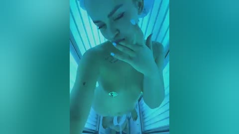 Media: Video of a topless woman with light skin and short, dyed hair, lying inside a tanning bed. She has small breasts and a silver belly button ring, with her hand covering her mouth.