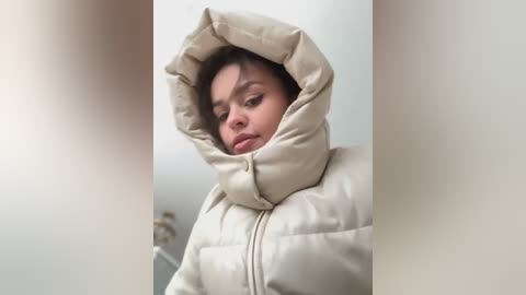 Media: A video of a woman with medium brown skin and brown hair, wearing a beige puffy coat with a hood, standing indoors with a neutral, contemplative expression.