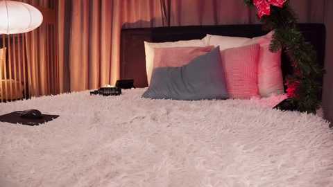 Media: Video of a cozy bedroom with a fluffy white shag rug, a black headboard, a bed with blue and pink pillows, a modern lamp, and festive greenery.