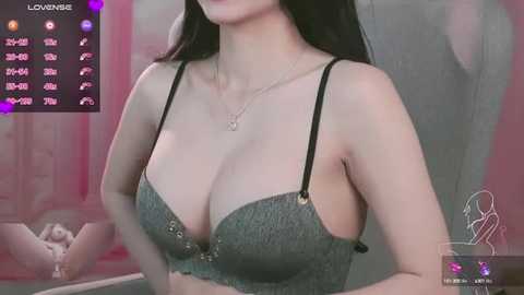 Media: Video of an East Asian woman with long black hair, wearing a grey lace bra, sitting on a bed. The background features a pink curtain and a digital display showing \"Lovense.\