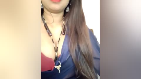 Media: Video of a woman with light brown skin, long dark hair, and red lipstick. She wears a deep red bra under a blue blouse with a chain necklace and dangling earrings.