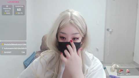 Media: A video of a light-skinned, blonde-haired woman with long hair, wearing a black mask, sitting in a white room with a gray door and a blue mat on the floor.