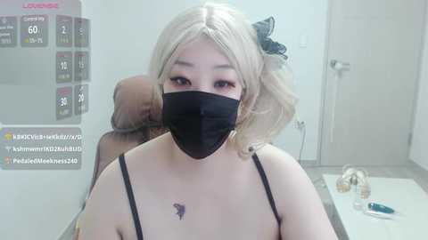 Media: Video of a young, fair-skinned woman with blonde pigtails, wearing a black face mask and black spaghetti straps top. She sits in a sterile medical setting, with a monitor displaying vital signs and a phone on the table.