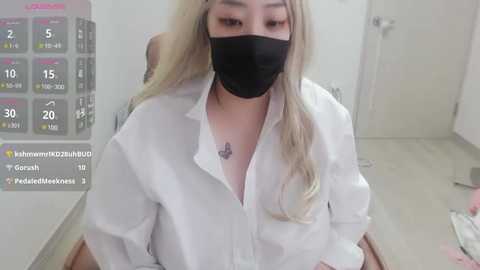 Media: A video of an Asian woman with long blonde hair and a black face mask, wearing a loose white shirt, standing in a sterile, white-walled room with a digital display showing \"Covid-19 Vaccine\" and her name.