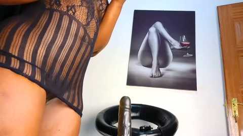 Media: Video of a woman in a sheer, black lace lingerie bodysuit, leaning on a black leather stool in a modern room with a minimalist decor.