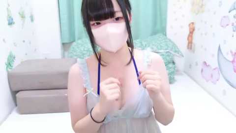 Media: Video of a young, fair-skinned Japanese woman with black hair, wearing a pastel-colored nightgown, face mask, and holding stethoscope, in a white room with light decor.