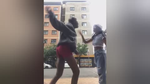 Media: Video of two young Black women, one in a black jacket and red shorts, the other in a striped shirt and blue jeans, mid-brawl on a sunlit urban street with multi-story beige and orange buildings in the background.