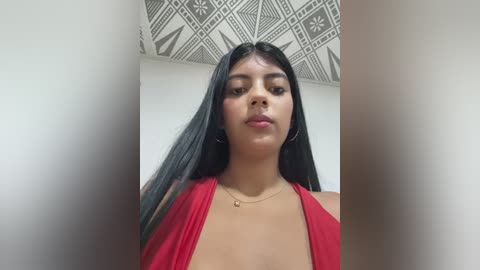 Media: Video of a young Latina woman with long black hair, wearing a red halter top, standing indoors with geometric patterned ceiling.