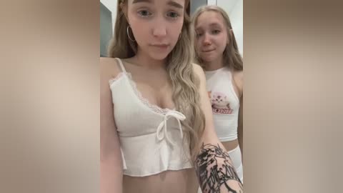 Media: Video of two young women with long blonde hair, wearing white crop tops, posing closely together. One has a tattoo on her arm. Background is blurred.