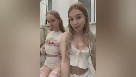 Media: Video of two young women in matching white crop tops and high-waisted shorts, sitting side by side, one smiling, the other looking down. Background shows a modern, brightly lit bathroom with white walls and a mirror.